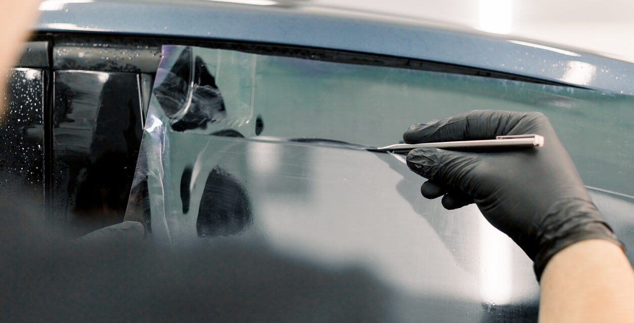 Cropped image of hands of worker in garage tinting a car window with tinted foil or film, holding special blade or knife to cut the film. Car detailing workshop, tinting windows.